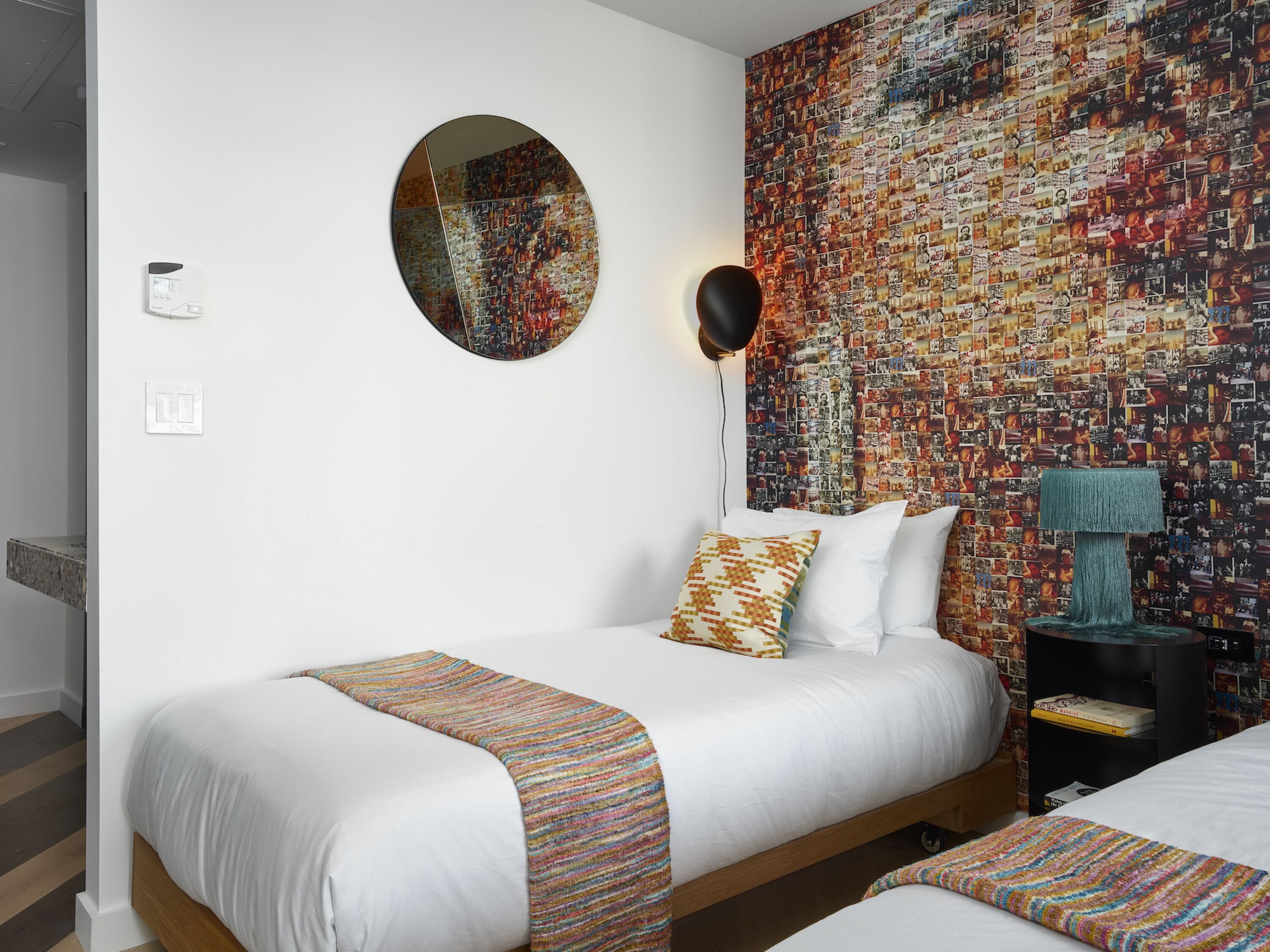 A hotel room with a bed, a round mirror, a wall covered with photos, a lamp, and a bookshelf.