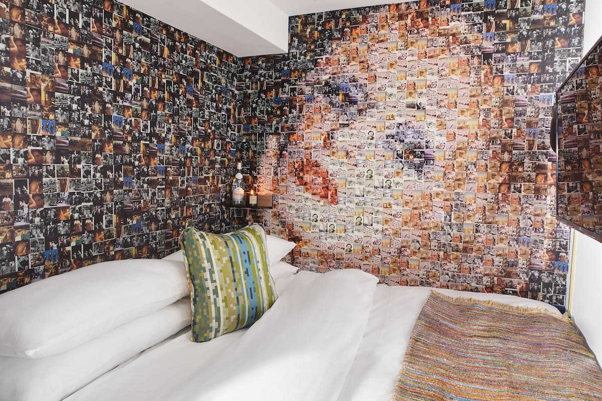 A bedroom with a wall covered in photographs, a bed with white linens, and a colorful pillow.