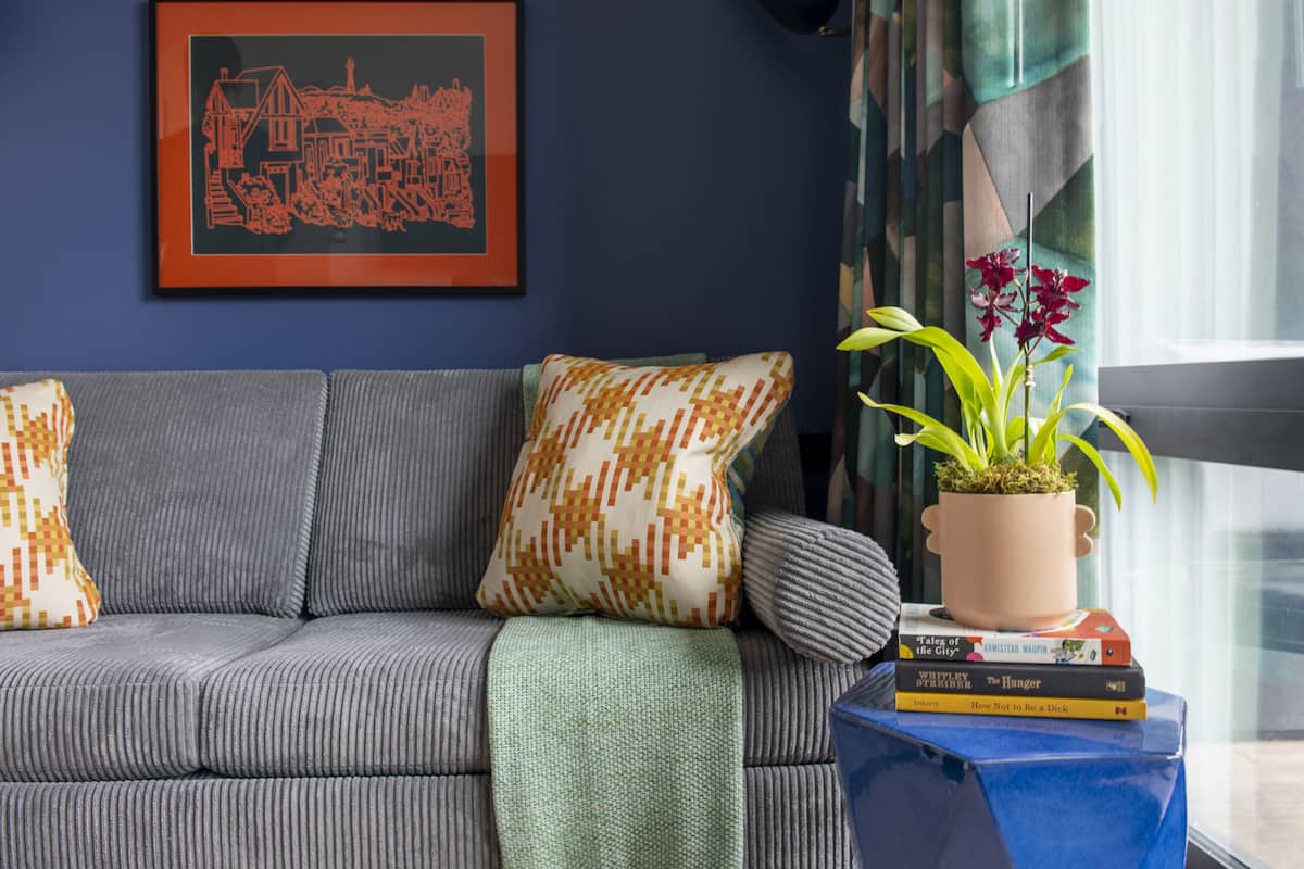 A modern living room with a grey sofa, patterned cushions, a framed artwork, a potted plant, and a stack of books.