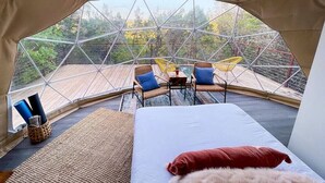 Een knus ingerichte tent met een bed, twee stoelen, een kleine tafel en uitzicht op het omliggende bos.