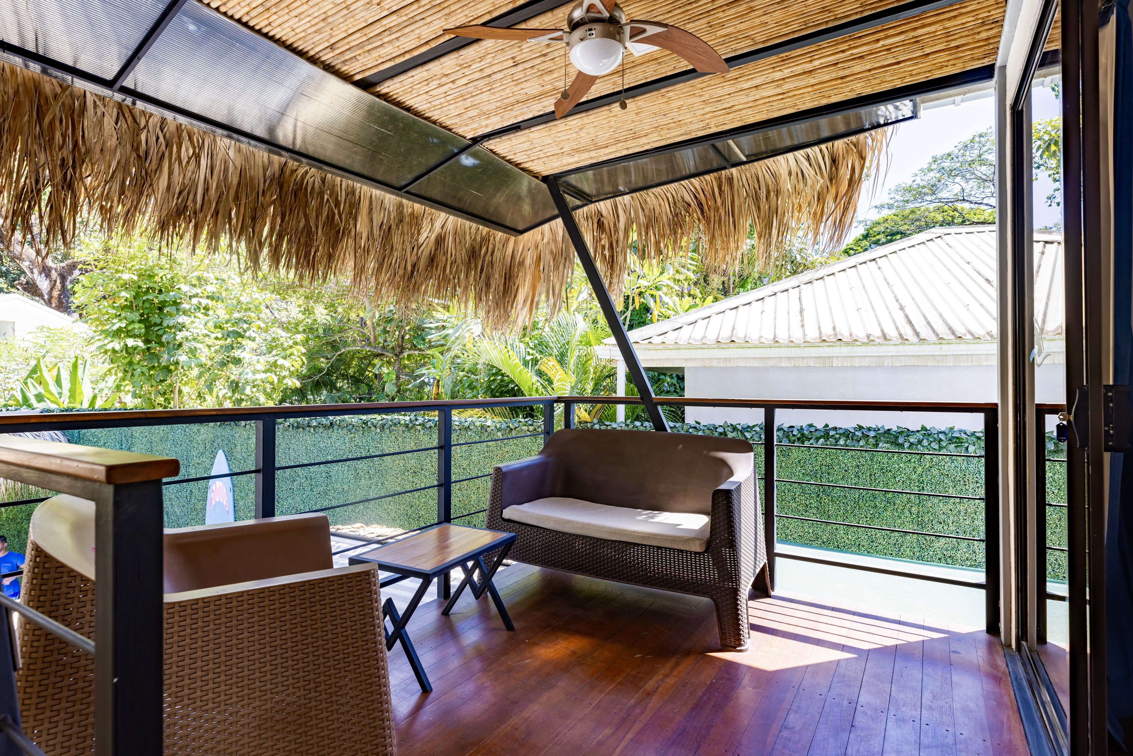 Uma área de estar coberta ao ar livre, com móveis de vime, um ventilador de teto e vista para o verde e um edifício ao fundo.