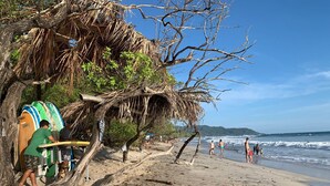 Una playa con tablas de surf, un letrero de alquiler de tablas y gente caminando por la orilla.