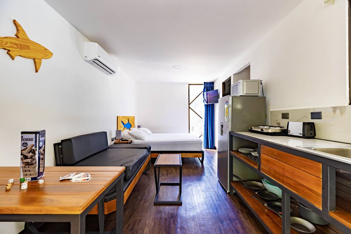 A compact hotel room with a bed, a small desk, a kitchenette, and a wooden wall decoration.