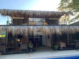 Un edificio de dos pisos con techo de paja, piscina y mobiliario exterior.