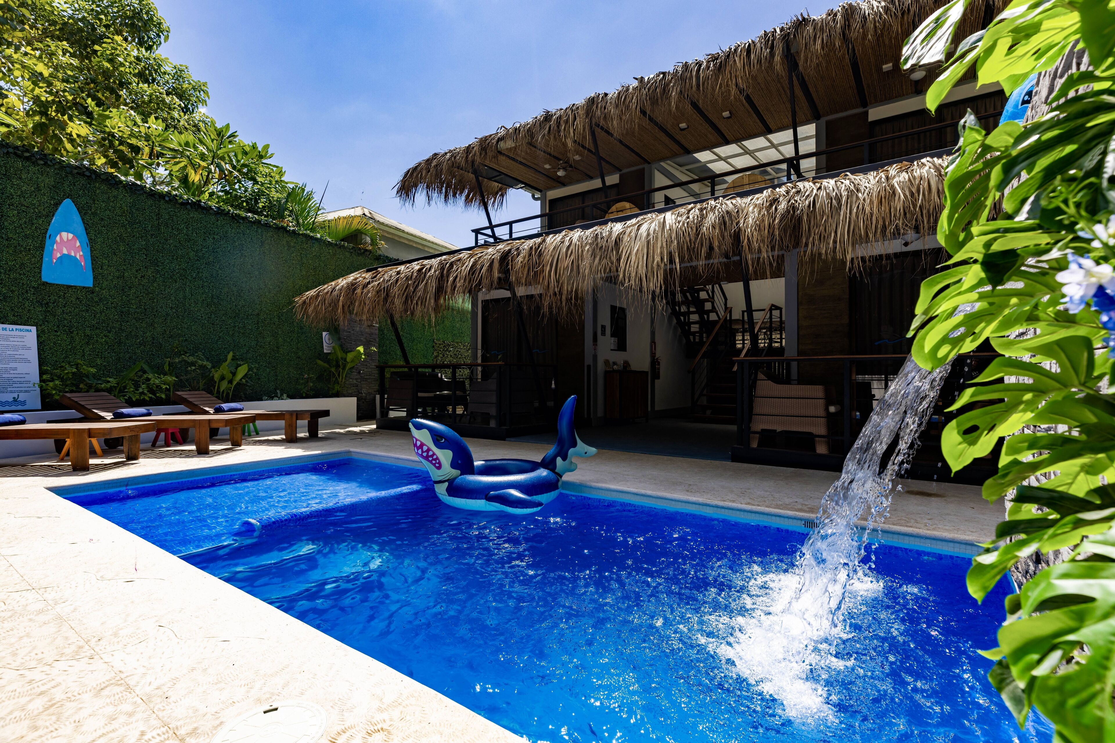 A swimming pool with a shark float, a thatched-roof structure, and a sign on the wall.