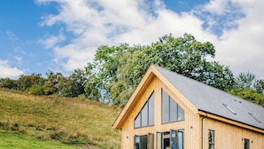 Una cabaña moderna de madera con ventanas amplias, techo inclinado y un paisaje de colina cubierta de césped.