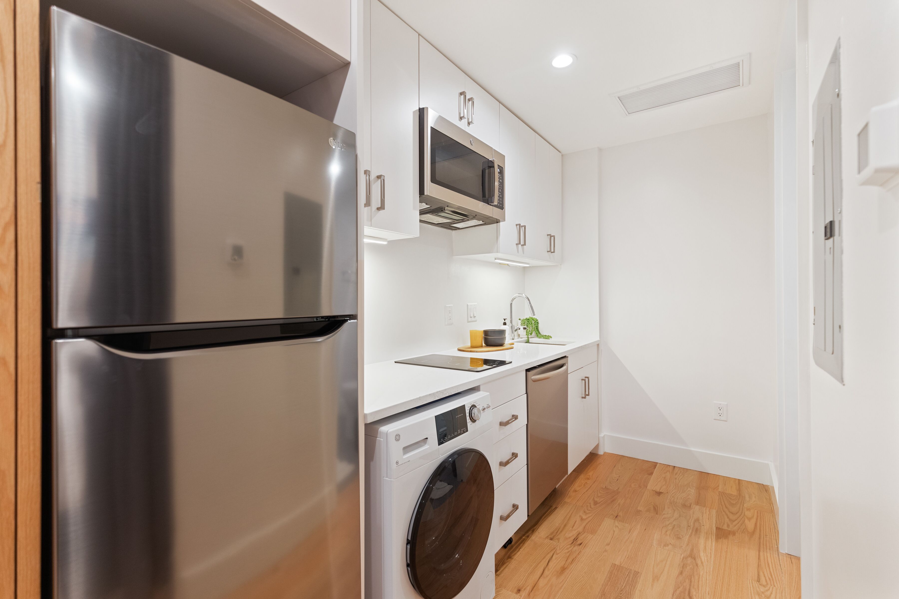 A compact kitchen with a stainless steel refrigerator, a microwave, a washing machine, and a dishwasher.