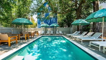 A swimming pool with lounge chairs and umbrellas, surrounded by trees and a blue building in the background.