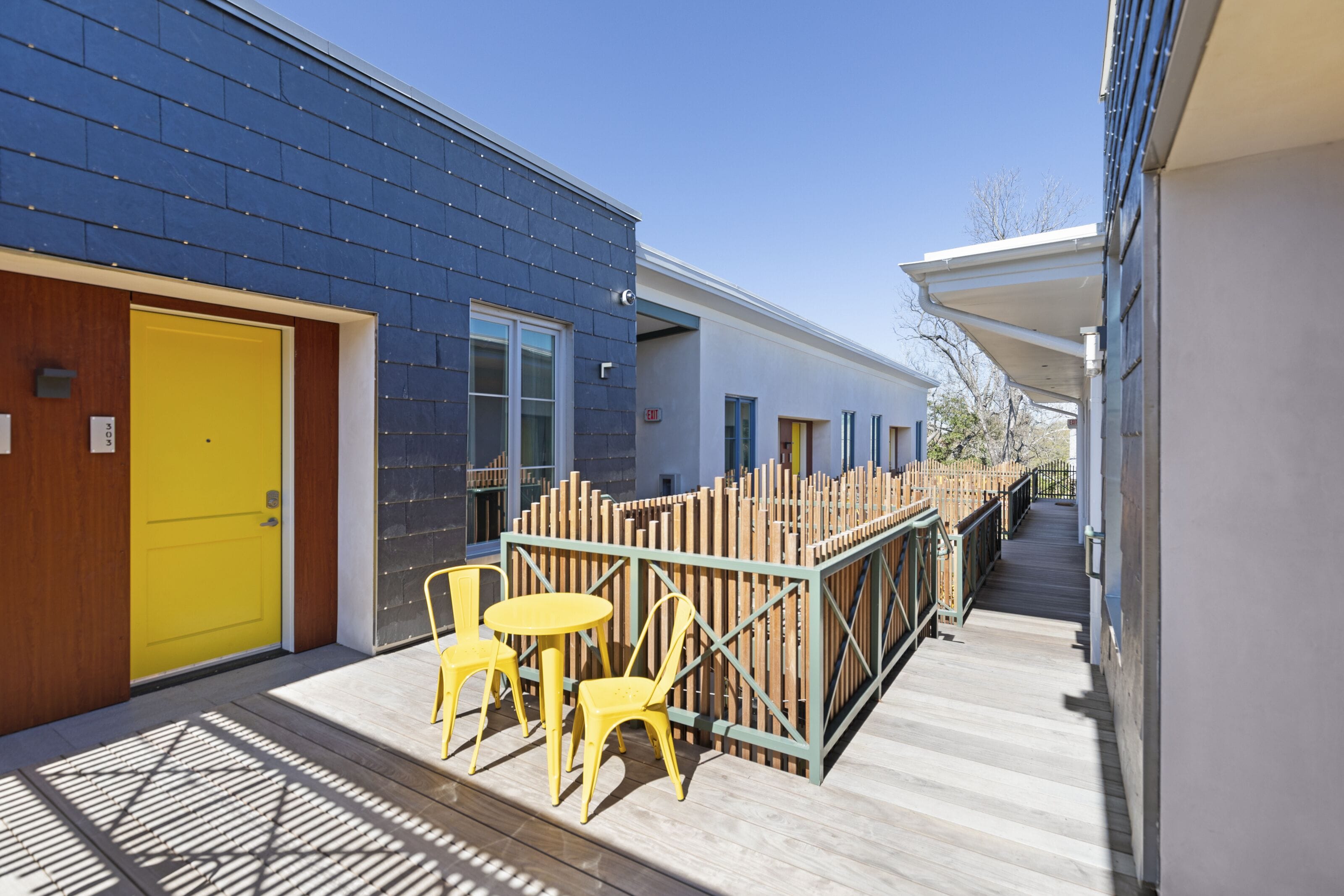 A modern building with a yellow door, a covered outdoor seating area with yellow chairs, and a wooden fence.