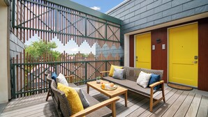 An outdoor seating area with wicker furniture and cushions, a wooden table with a tray of items, and a yellow door in the background. - The Iris Charleston (Charleston)