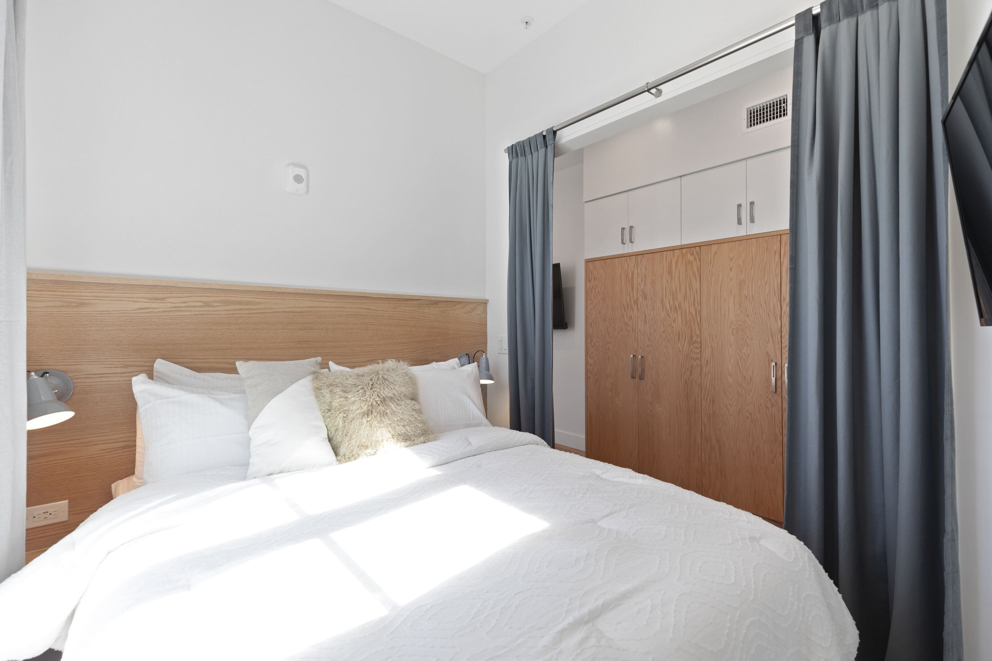 A modern bedroom with a wooden headboard, a large bed with white bedding, a nightstand with a lamp, and a wardrobe with a mirror.