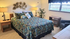A bedroom with a bed, two bedside tables, a nightstand, a chair, a window, and a plant. - 108 - 2 BR Poolside Great View (New Smyrna Beach)