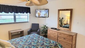 A bedroom with a bed, a chair, a dresser, a television, and a window with a view. - 108 - 2 BR Poolside Great View (New Smyrna Beach)