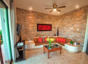 A living room with a stone wall, a ceiling fan, a red abstract painting, a sectional sofa with red cushions, a coffee table with magazines, and a small potted plant.