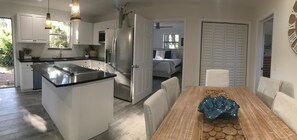 Eine moderne Küche mit weißen Schränken, einer schwarzen Arbeitsplatte und einem Edelstahl-Kühlschrank. Es gibt einen Essbereich mit einem Holztisch und Stühlen, und ein Schlafzimmer ist durch eine offene Tür sichtbar.