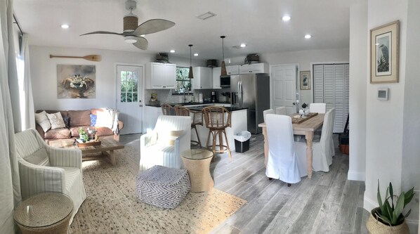 Ein großzügiger Wohnbereich mit einer Couch, Sesseln, einem Couchtisch und einem Esstisch mit Stühlen. Es gibt eine Küche mit Kühlschrank, Herd und Schränken.