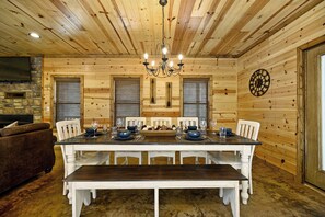 Un comedor de madera con una gran mesa dispuesta para seis comensales, flanqueada por dos ventanas con persianas, una chimenea de piedra y un techo de madera con un candelabro.