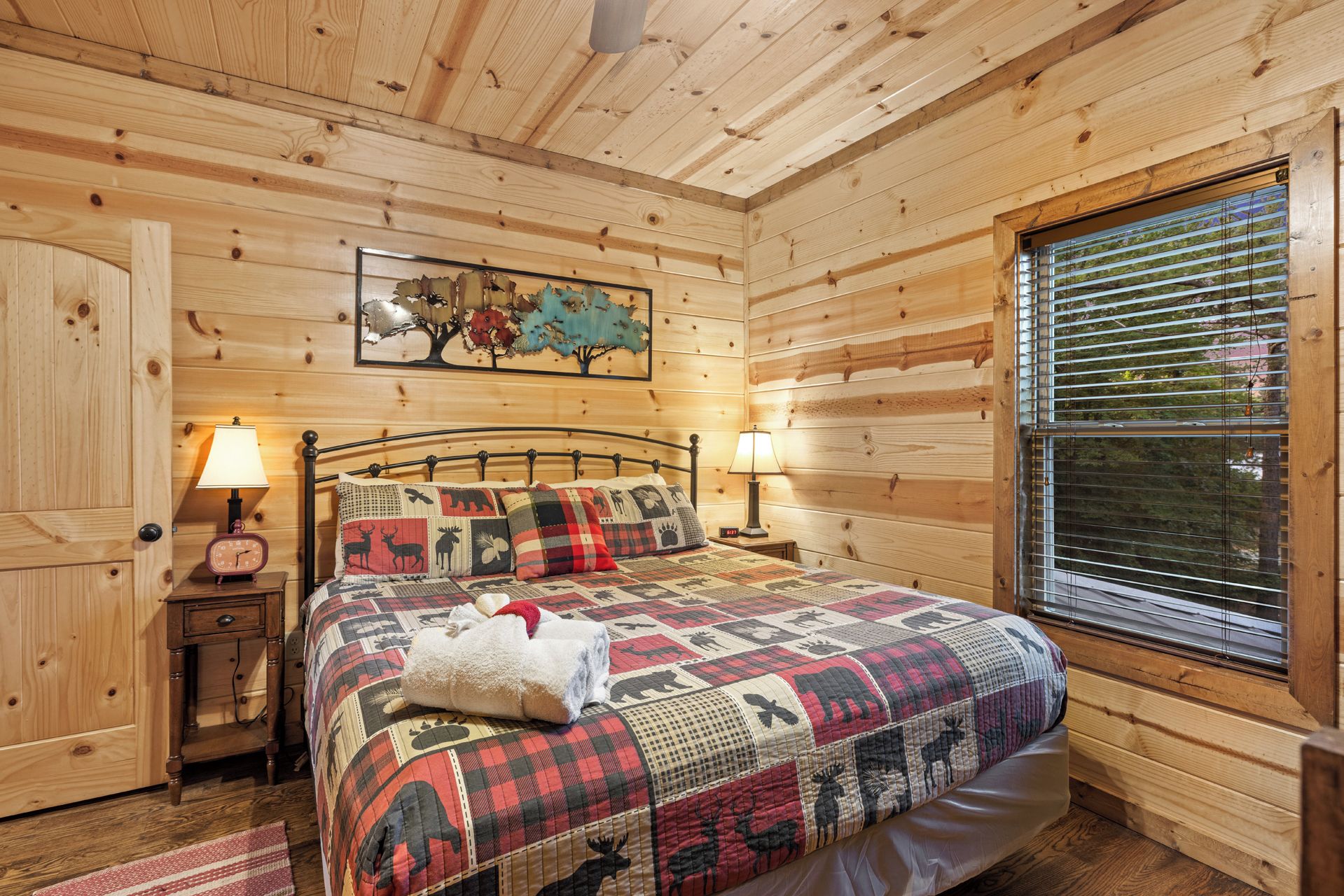A wooden cabin bedroom with a bed, two bedside lamps, a clock, a window with blinds, and a wall painting.