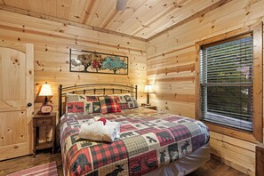 A wooden cabin bedroom with a bed, two bedside lamps, a clock, a window with blinds, and a wall painting.
