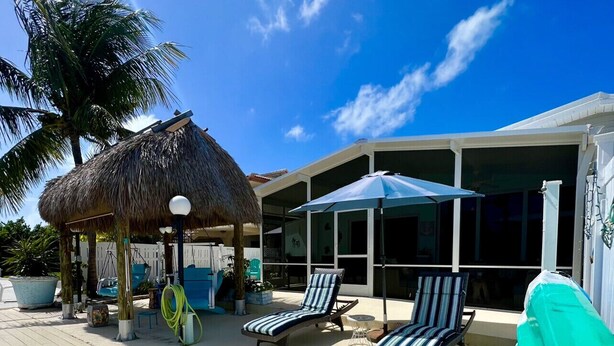 A wooden deck with lounge chairs and a thatched-roof structure, likely a cabana, adjacent to a building with large windows and a covered area with umbrellas.