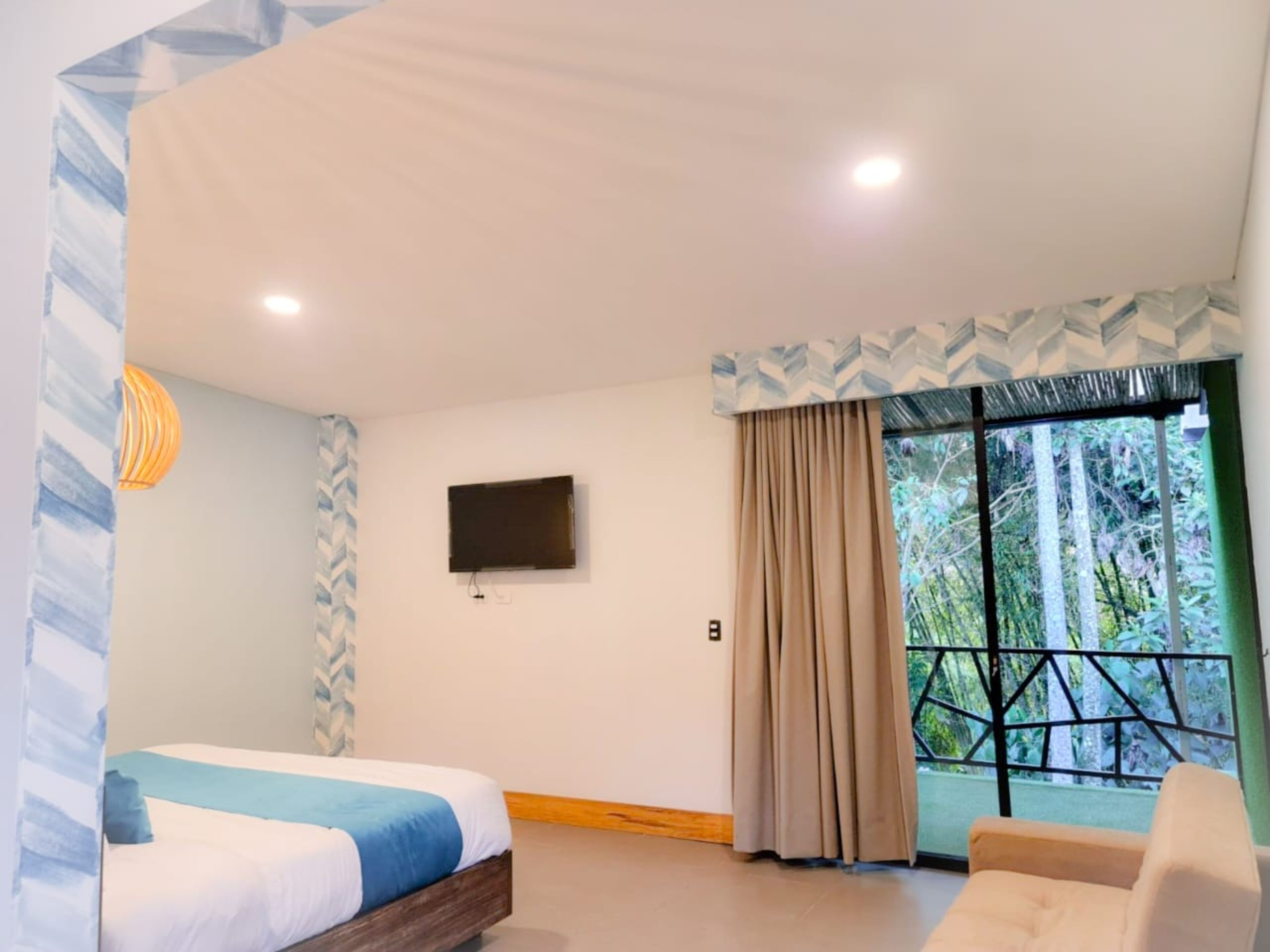 A hotel room with a bed, a TV mounted on the wall, a window with curtains, and a view of greenery outside.