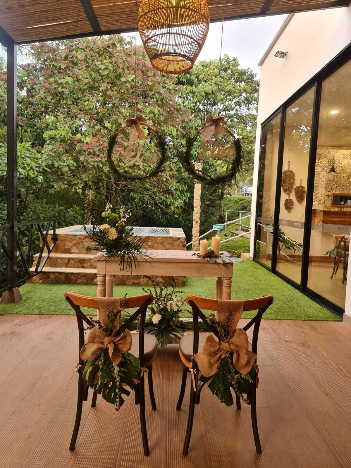 An outdoor dining area with a wooden table and chairs, surrounded by greenery and a view of trees.