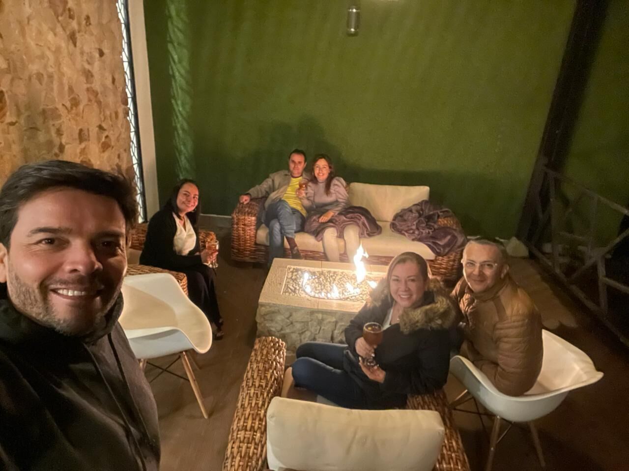 Un grupo de personas sentadas alrededor de una mesa con una vela encendida, en una habitación con muebles de mimbre y una pared verde.