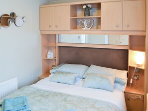 Ein ordentlich bezogenes Bett mit einem blumigen Überwurf und Kissen, ein Nachttisch mit Lampe und ein Wandregal mit Dekoration.