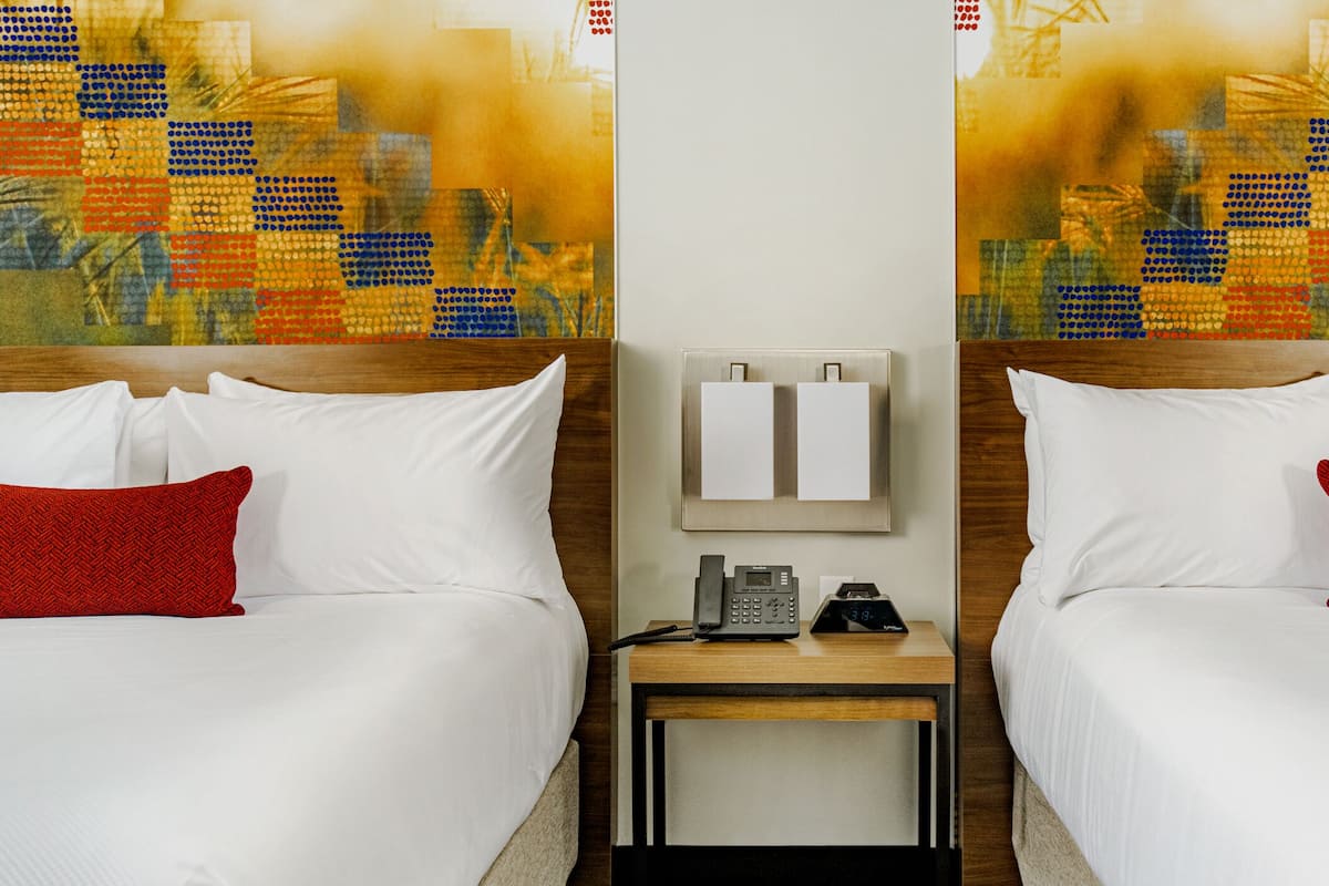 Een hotelkamer met twee bedden, elk voorzien van witte beddengoed en rode kussens, een nachtkastje met een telefoon en een digitale klok, en een wandlamp.