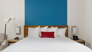 Hotelkamer met een groot bed, een houten hoofdbord, wit beddengoed, een rood sierkussen, een lamp, een stoel en een nachtkastje met een telefoon.