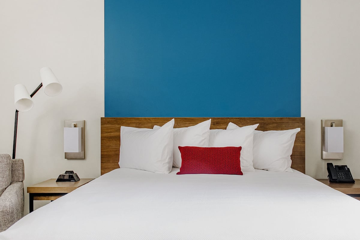Hotelkamer met een groot bed, een houten hoofdbord, wit beddengoed, een rood sierkussen, een lamp, een stoel en een nachtkastje met een telefoon.