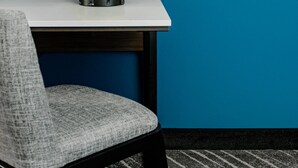 Een stoel met een zitkussen van grijze stof en een zwart frame, geplaatst voor een tafel met een wit blad en een gele lamp met een witte lampscherm.