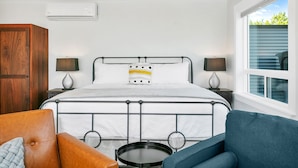 Une chambre à coucher avec un lit en métal, deux tables de chevet avec des lampes, une armoire en bois et une vue sur l’extérieur par une fenêtre.