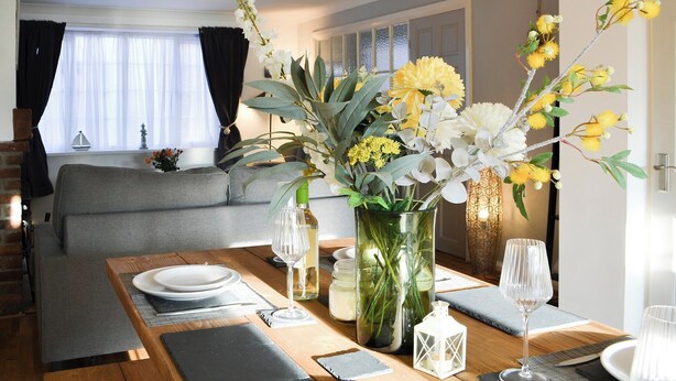 Un comedor con una mesa de madera preparada para dos personas, un florero con flores y vista a una sala de estar con sofá y cortinas.