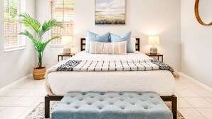 Un dormitorio con cama, lámparas de noche, un ventanal grande, una planta en maceta y un cuadro en la pared.