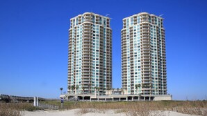 Deux immeubles de grande hauteur, de forme cylindrique, dotés de balcons, situés près d’une plage de sable où la végétation est clairsemée.