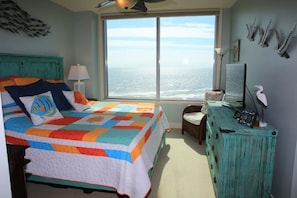 A bedroom with a large window, a bed with a colorful quilt, a dresser, and wall art. - Palisade Palms, Cheerful, Modern Condo with Private Patio and Beachfront Access (Galveston)