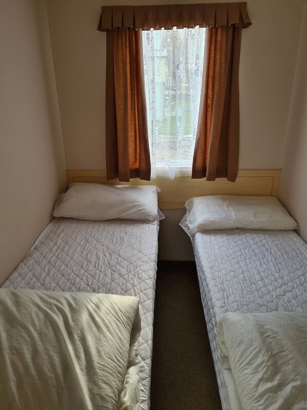 A room with two single beds, a window with brown curtains, and a view of greenery outside. - Happy Days Chapel st Leonards 3bed 8berth (chapel st leonards)