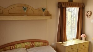 Ein ordentlich bezogenes Bett mit weißen Leinen, ein Holzkopfschild mit verzierten Herzformen, ein Nachttisch mit Blumen in einer Vase und ein Fenster mit Vorhängen.