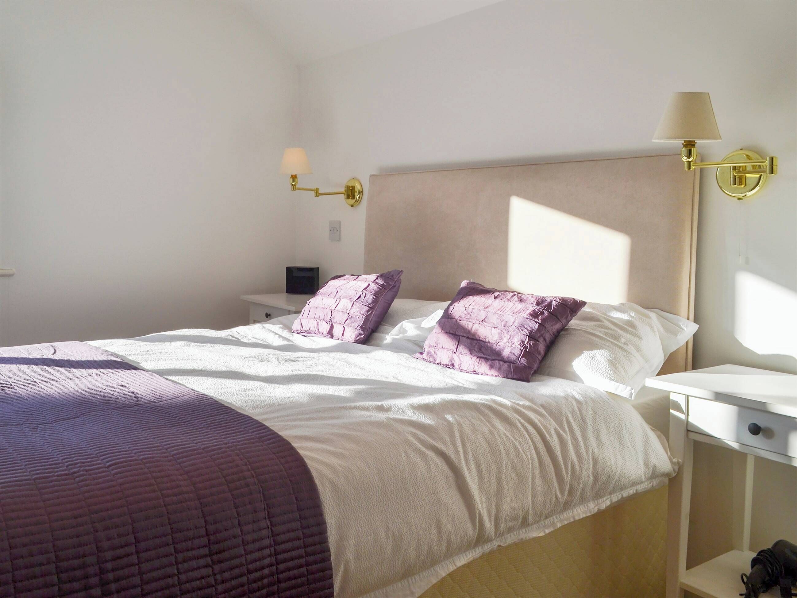A neatly made bed with a white and purple quilt, a bedside table with a lamp, and a nightstand with a laptop.