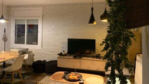 A modern living room with a white brick wall, a flat-screen TV, wooden furniture, and hanging plants. - Apartment in guarded residence overlooking swimming pool (Platja d'Aro)