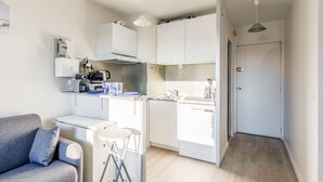 Una cocina compacta con armarios blancos, horno empotrado y un lavamanos. Hay un pequeño comedor con una mesa blanca y dos taburetes. Un sofá gris con un cojín está apoyado contra la pared.