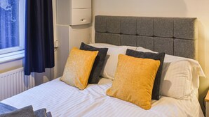 A neatly made bed with white linens and two mustard-colored pillows, a grey upholstered headboard, and a small bedside table with a lamp. - A delightful haven for walkers and cyclists. (Skipton)