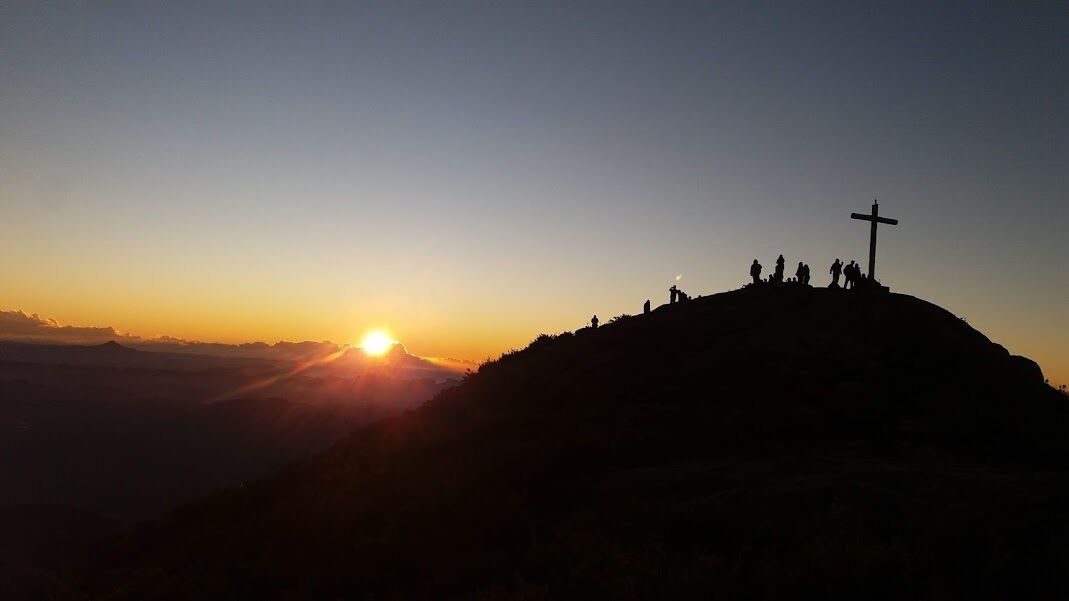 A silhouette of people on a hill with a cross, and the sun setting in the background.