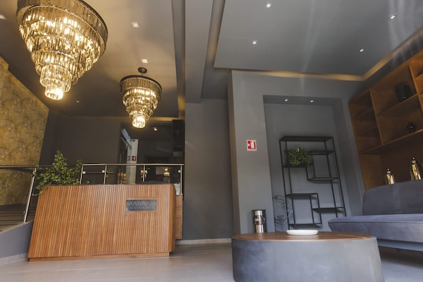 A modern reception area with a wooden reception desk, two large chandeliers, a grey sofa, and a small table with a white object on it.