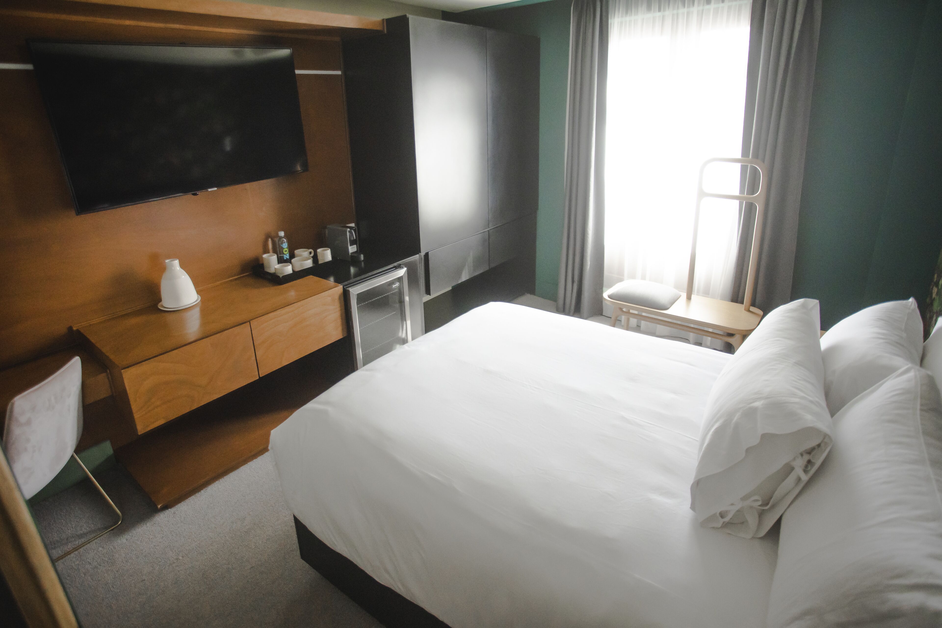 A hotel room with a bed, a TV mounted on the wall, a desk with a chair, and a window with curtains.