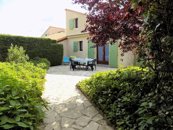 Un chemin de pierres mène à une table dressée pour deux personnes, dans un coin jardin, avec un bâtiment visible en arrière-plan.