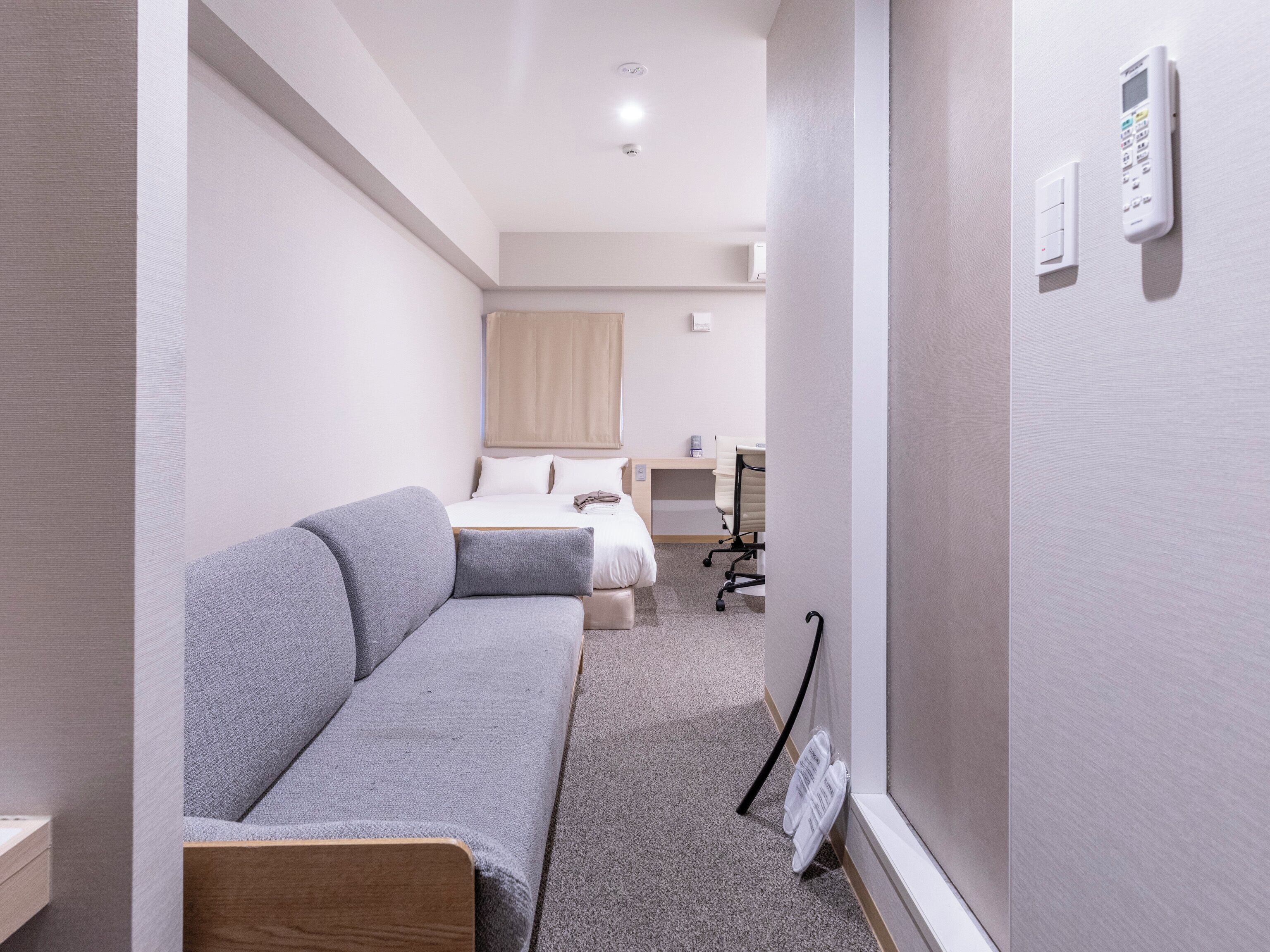 Quarto de hotel moderno com um sofá cinzento, uma cama com roupa de cama branca, uma escrivaninha com uma cadeira e um telefone fixado na parede.