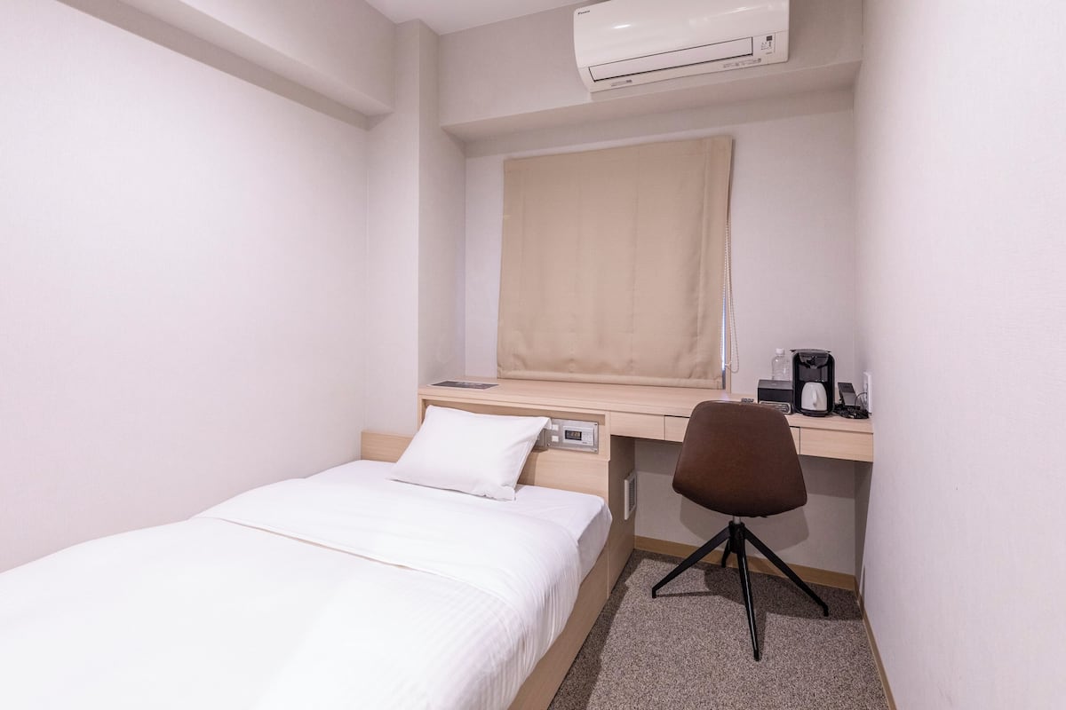 Una habitación pequeña y limpia con cama, escritorio, silla, aire acondicionado y ventana con persiana.