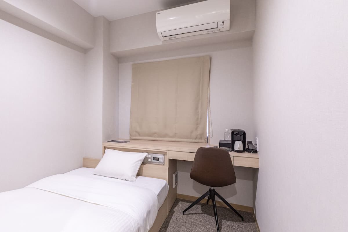 A hotel room with a single bed, a desk with a chair, a coffee maker, and an air conditioner.